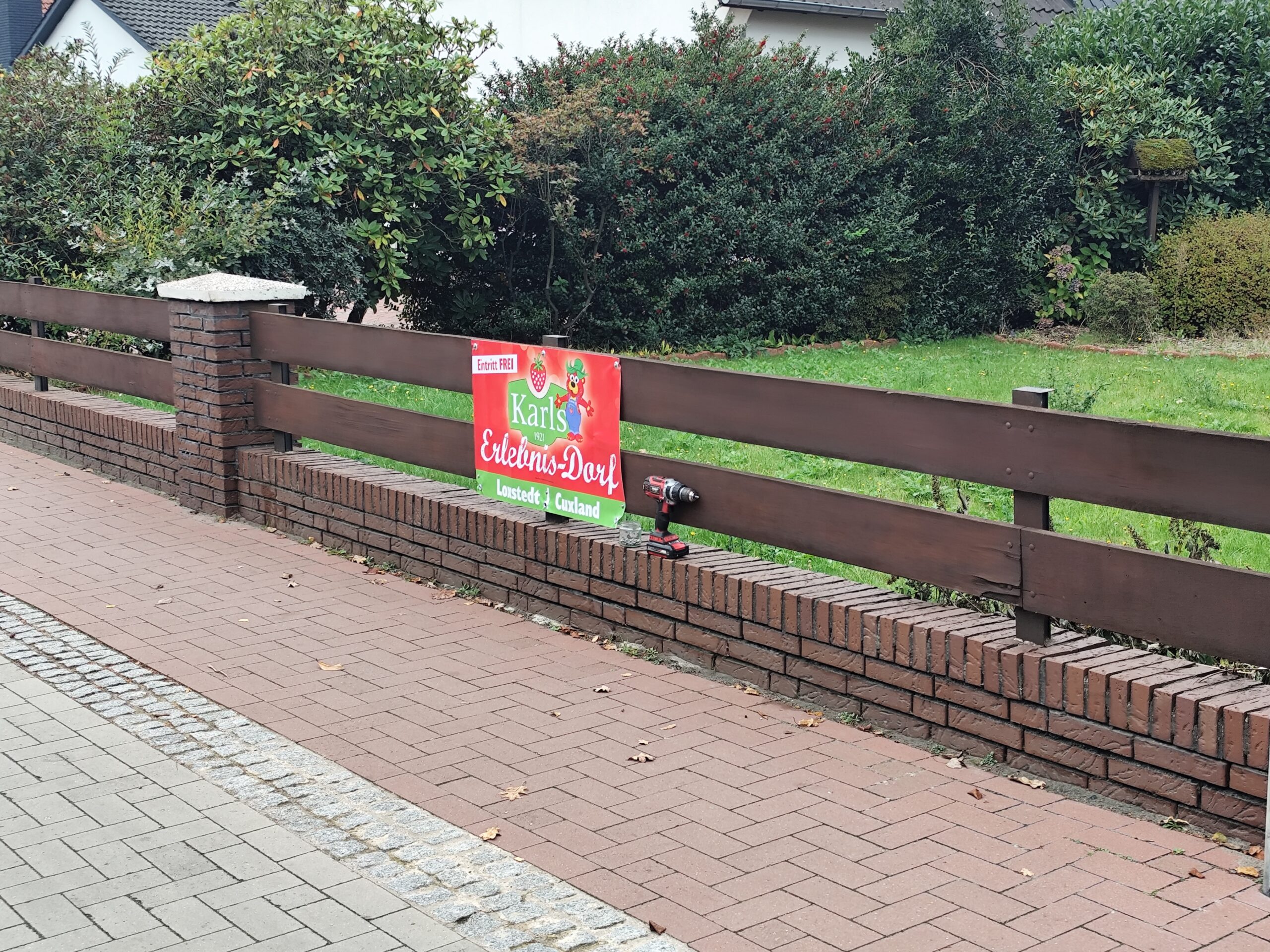 Das Foto zeigt einen Zaun an der Straße, der durch breite lange Bretter zwischen Steinposten steht. Angebracht ist eine Werbung für Karls Erlebnisdörfer